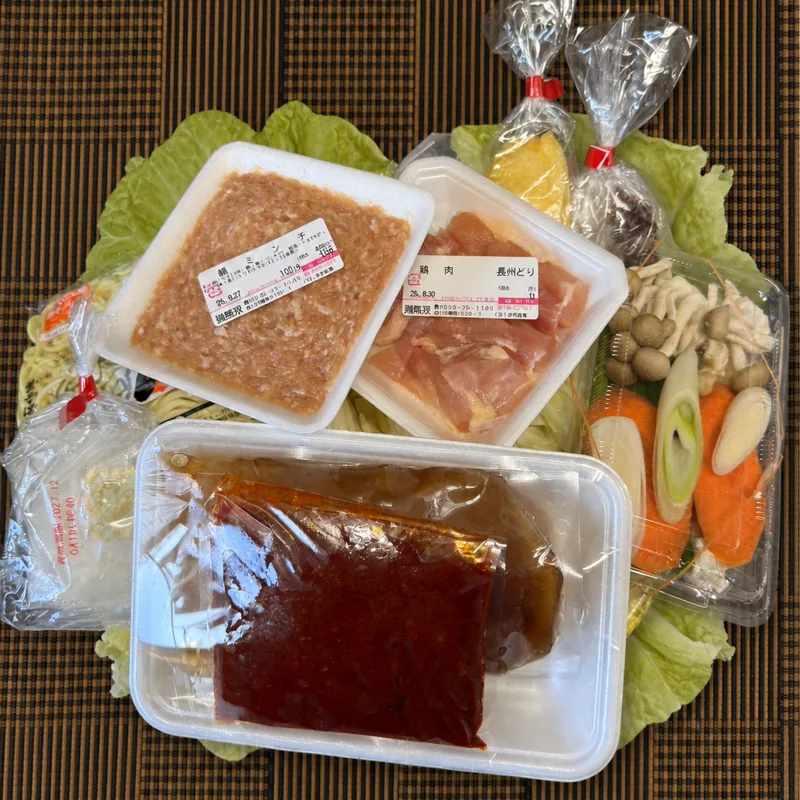 辛鍋セット一式。ござの上に敷いたキャベツと白菜に、ピリ辛スープ、鍋ミンチ、鍋肉（長州どり）、カット野菜、〆のちゃんぽん麺を並べたところ。